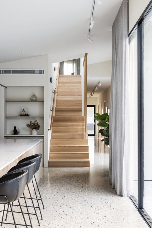 Bright open-plan residential interior featuring a timber staircase, terrazzo flooring, a marble-look kitchen island with dark bar stools, built-in display shelving, floor-to-ceiling glazing with sheer curtains, and track lighting. The view is primarily toward the stairway and adjoining hallway.