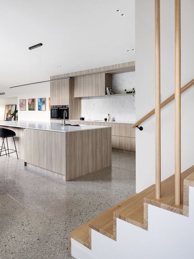 Bright contemporary open-plan kitchen viewed beside a timber staircase. The kitchen features light timber cabinetry, a large island with a pale stone benchtop, integrated black appliances, a white marble-look splashback, and a polished terrazzo floor. The staircase has white risers, timber treads, and vertical timber balustrade posts with a simple handrail. Minimal furnishings and neutral finishes create an airy, refined interior.