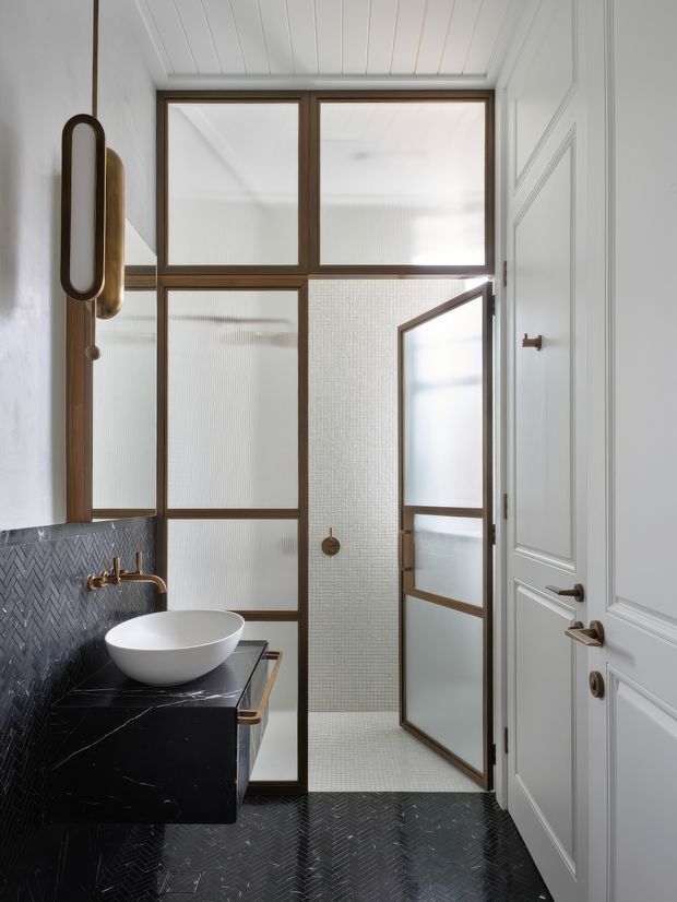 A refined bathroom featuring a black marble floating vanity with a white vessel basin and brass tapware. Dark herringbone wall and floor tiles contrast with white paneled walls and ceiling. A framed fluted-glass shower enclosure with a hinged door occupies the rear of the room, while a large wall mirror and decorative wall lighting add depth. The overall scheme is compact, elegant, and high-contrast.
