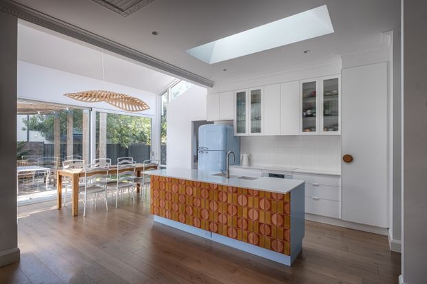 Bright open-plan kitchen and dining area with white built-in cabinetry, glazed display cupboards, a pale stone island benchtop, patterned orange and mustard tiled island front, timber flooring, and a vintage-style blue refrigerator. A large skylight brings in natural light, while full-height glazing connects the dining area to an outdoor space. The room combines classic architectural detailing with colourful retro-inspired joinery.