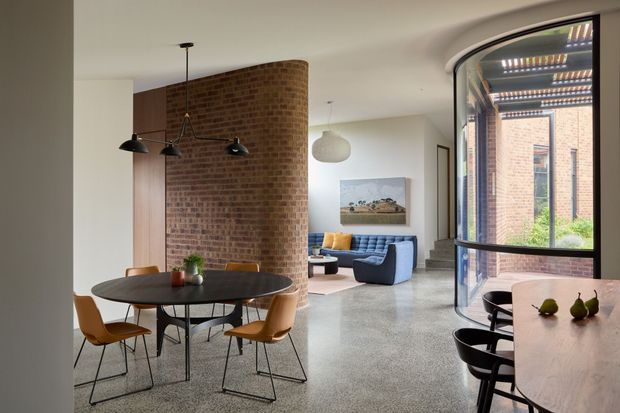 Open-plan contemporary dining and living area with a round dark dining table and sculptural pendant lights in the foreground. A curved exposed-brick partition separates the dining zone from a lounge furnished with a blue sofa and armchair. Large curved glazing opens views to a brick courtyard and pergola, while terrazzo flooring runs throughout the space.