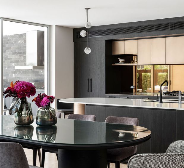 Contemporary open-plan kitchen and dining area with dark textured cabinetry, a pale stone island, integrated appliances, black-framed glazing, and a glossy oval dining table with upholstered chairs. Three spherical pendant lights hang above the island, while a dramatic dark stone wall is visible through the glazing.