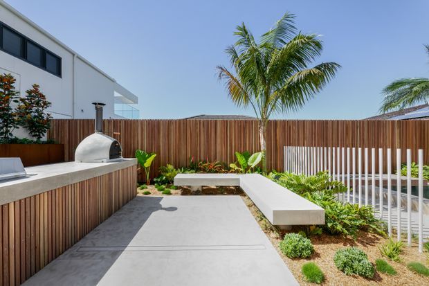 A contemporary outdoor entertaining area with a built-in curved pizza oven, concrete bench and dining surfaces, vertical timber privacy fencing, white metal balustrade, concrete paving, and tropical planting including palm trees. A modern white building is visible beyond the courtyard.