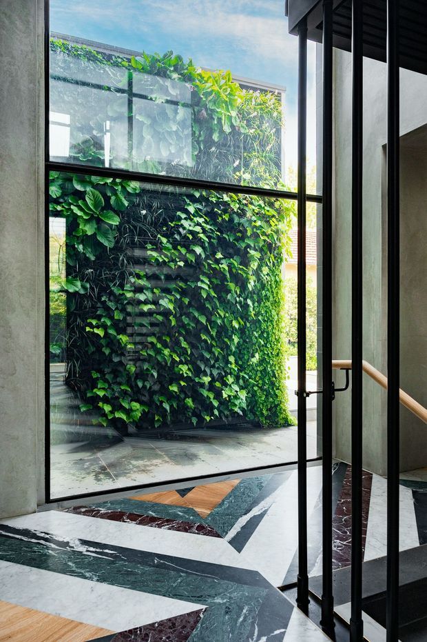 A dramatic interior view toward a lush vertical garden courtyard through large black-framed glazing. The space features a geometric stone and marble floor in white, dark green, burgundy and timber-toned sections, raw grey plaster walls, and a black-framed glazed stair enclosure with a timber handrail. Strong daylight, greenery and bold material contrasts create a refined, biophilic atmosphere.