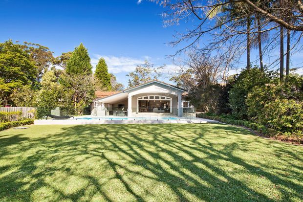 Single-storey suburban house viewed from a large landscaped lawn, with a central gable-roofed rear façade, broad covered outdoor entertaining area, extensive glazing, and a swimming pool immediately in front. Mature trees and hedges surround the property.