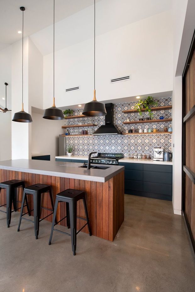 A contemporary kitchen with a large island and integrated seating, featuring a richly patterned tile splashback, dark lower cabinetry, timber-clad island panels, and a concrete-look floor. Three black pendant lights hang over the island, while open timber shelves display plants and kitchenware. The kitchen has a high, bright ceiling and a warm, refined material palette.