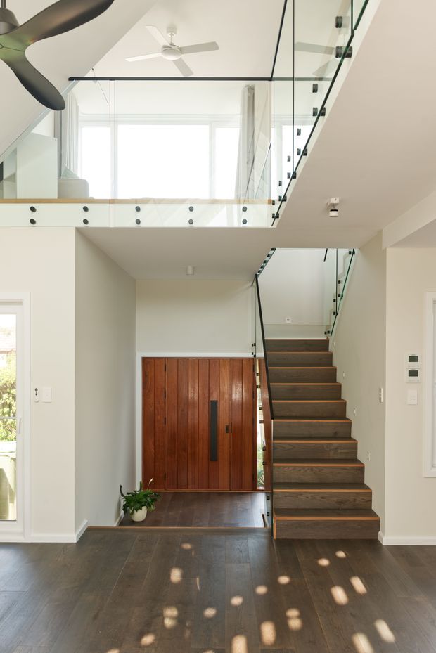 Bright contemporary double-height entry hall with a floating timber staircase, transparent glass balustrades, dark timber flooring, and a mezzanine-level opening. A large timber entry door sits beneath the stairs, while ceiling fans and generous windows bring in natural light.