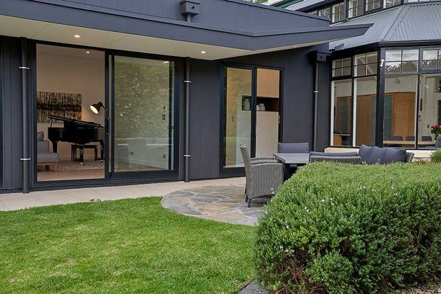 Exterior view of a dark-clad contemporary home opening onto a landscaped courtyard and outdoor seating area. Large glazed doors reveal a piano room and a light-filled bathroom or living space inside. The courtyard includes a paved stone terrace, wicker-style lounge furniture, lawn, and manicured shrubs. A pitched corrugated metal roof and contrasting older-style glazed structure are visible at right.