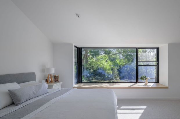 Bright, minimalist bedroom with a large black-framed window overlooking leafy greenery. The room features a low-profile upholstered bed with pale grey bedding, white walls, light carpet, a small white bedside table, and a timber-toned window sill. Natural light creates a calm, airy atmosphere.