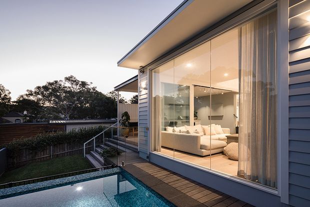 Exterior view of a contemporary residential extension at dusk, with blue-grey horizontal cladding, broad floor-to-ceiling glazed doors, and a timber deck overlooking a swimming pool and landscaped backyard. Through the glazing, a bright living room with a large cream sectional sofa and neutral furnishings is visible.