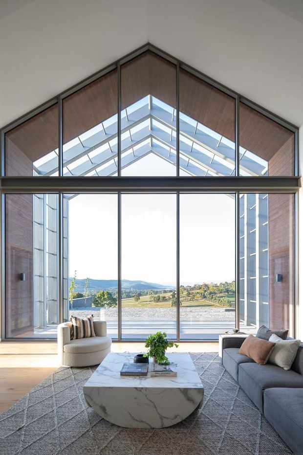 A bright, refined living room centered on a dramatic full-height glazed gable wall. The room features a high pitched ceiling with exposed timber lining, dark slender window frames, extensive clerestory-style glazing, and views across a rural rolling landscape. A grey sectional sofa and rounded cream armchair sit around a large sculptural marble coffee table on a textured grey rug.