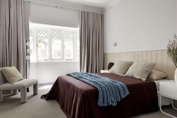 A refined, softly lit bedroom with a large upholstered bed dressed in deep burgundy bedding, layered neutral pillows, and a chunky blue-grey throw. The room features full-height taupe curtains, a pale carpet, a white-framed bay window with diamond-pattern glazing, and a two-tone wall treatment with vertical ribbed panelling above a beige lower section. A sculptural upholstered chair and minimalist white bedside table complete the calm, contemporary interior.