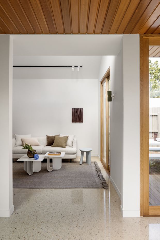A bright, refined living room viewed from an open entry or hallway. The room features a white upholstered sofa, sculptural white coffee tables, a small side table, a neutral textured rug, and restrained artwork. White plaster walls contrast with a warm timber-lined ceiling and timber-framed glazing and doors opening toward a deck. The foreground has a polished light terrazzo floor, while the space is illuminated by natural light and minimalist track lighting.