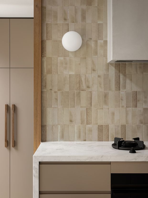 Close-up view of a refined contemporary kitchen, featuring a vertically stacked beige and taupe ceramic tile splashback, pale veined stone benchtop, matte warm-neutral cabinetry with timber handles, black gas cooktop, and a simple spherical wall light. The composition highlights the fixed joinery and material detailing.