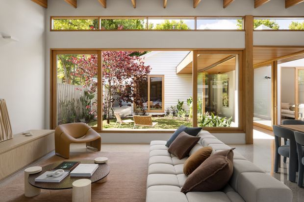 A bright contemporary living room opening onto a landscaped courtyard through expansive timber-framed glazing. The room features exposed timber ceiling beams, warm timber window frames and joinery, a large light-coloured sectional sofa, sculptural armchair, layered coffee tables, and a dining area visible at right. The courtyard and adjoining white-clad structure are visible beyond the glazing.