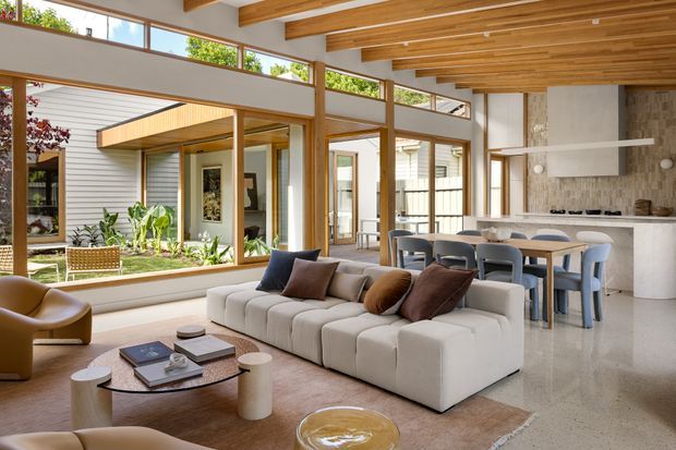 A bright, open-plan living and dining area with a contemporary architectural design. The room features extensive floor-to-ceiling glazing overlooking a planted courtyard, exposed warm timber ceiling beams and boards, pale horizontal wall cladding, polished light terrazzo or concrete flooring, and a stone kitchen island with integrated cabinetry. A large cream sectional sofa, sculptural lounge chairs, a timber dining table, and muted cushions create a warm, refined interior.