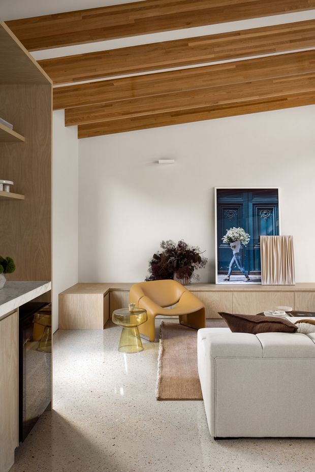 A refined contemporary living room with a warm timber-lined pitched ceiling, pale built-in cabinetry, terrazzo flooring, and a large cream sectional sofa. A sculptural mustard-yellow lounge chair, glass side table, artwork, dried floral arrangement, and textured rug add colour and character. The room includes a built-in media or display wall and a partial kitchen or joinery area at left.
