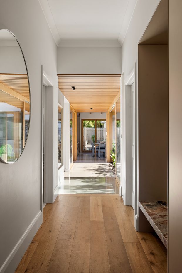 A bright, refined residential hallway viewed toward a dining area at the far end. The corridor has warm timber flooring, white walls with traditional-style cornice detailing, white door surrounds, full-height glazed openings, and a warm timber slatted ceiling continuing into the rear zone. A large rounded wall mirror is visible on the left, while built-in shelving with a dark stone or marble-like surface appears on the right. Sunlight creates patterned shadows across the pale floor, and greenery is visible through the glazing.