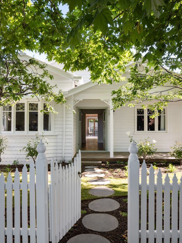A bright white weatherboard house viewed from the front garden through an open white picket gate. The facade features a central recessed entry with decorative timber trim, multi-pane windows, pitched roof forms, and a stepping-stone path through landscaped garden beds. Mature leafy trees frame the upper portion of the image.