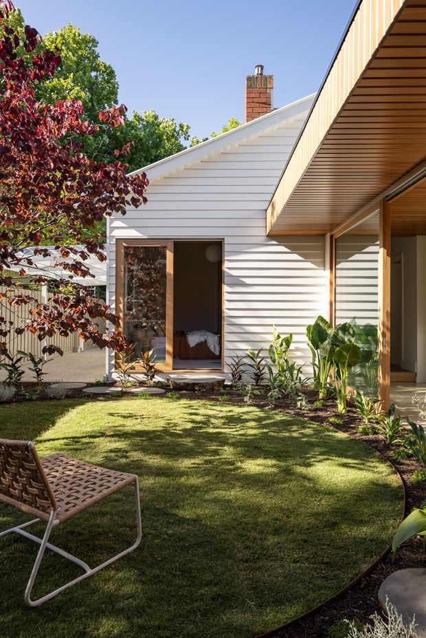 A bright exterior view of a contemporary single-storey home arranged around a landscaped courtyard garden. The white horizontal weatherboard facade has a pitched gable roof and exposed brick chimney, while a timber-lined covered terrace projects along the right side with large glazed openings. A bedroom is visible through an open doorway, and the courtyard includes lawn, garden beds, mature planting, and an outdoor chair.