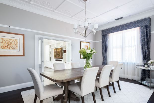 Bright, refined formal dining room with a substantial dark wood dining table surrounded by upholstered dining chairs. The room features pale grey walls, white architectural trim, ornate decorative ceiling mouldings, a traditional chandelier, patterned navy drapery, sheer curtains, framed artwork, and light tiled flooring. A wide opening connects the dining room to a bright adjacent interior space.