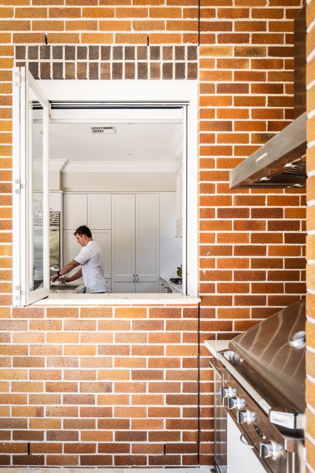 A bright architectural view of an outdoor barbecue kitchen against a red brick wall, with a stainless-steel built-in grill in the foreground and an open serving window looking into a white, traditionally detailed interior kitchen. The image emphasizes the brick facade, outdoor cooking equipment, and connection between indoor and outdoor spaces.