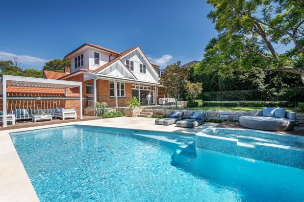 Bright exterior view of a substantial multi-level brick home with white trim, steep gabled roof sections and terracotta roof tiles. The landscaped backyard is dominated by a large swimming pool with an integrated raised spa or shallow ledge, paved surrounds, outdoor lounge seating, and a low stone retaining wall. Mature trees and hedging frame the property.