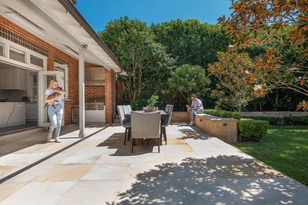 Sunny residential outdoor entertaining area adjoining a brick house, with a paved terrace, outdoor dining table and chairs, built-in barbecue bench, and a partially visible kitchen through open doors. Mature garden planting surrounds the patio.