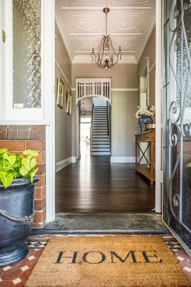 View through an open front door into a long, refined residential hallway. The entry features dark timber flooring, muted sage-grey walls with white trim, an ornate pressed-tin ceiling with decorative medallions, a traditional chandelier, framed artwork, a timber console table, and a staircase beyond. The threshold has patterned geometric tile flooring, a brick exterior wall, decorative leadlight-style security glazing, and a welcome mat.