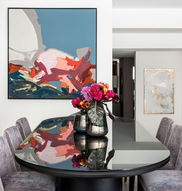 Contemporary dining area with a glossy black oval dining table, upholstered dining chairs, and a sculptural floral arrangement. The reflective tabletop mirrors the colourful abstract artwork and flowers, while white walls, a dark doorway, and streamlined built-in architectural elements create a refined, gallery-like setting.