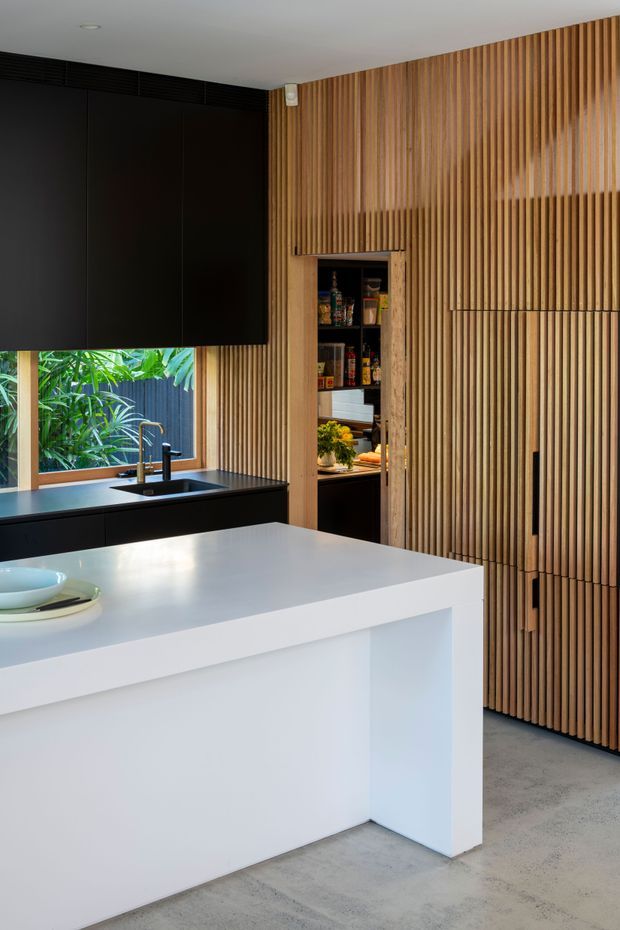Contemporary kitchen featuring a large white island with integrated open leg space, a dark benchtop and cabinetry, black wall-mounted cabinetry, and a full-height vertical timber batten feature wall concealing storage doors. A compact walk-in pantry is visible through an open door, with abundant greenery seen outside the window. The room has polished concrete flooring and a clean, high-contrast material palette.