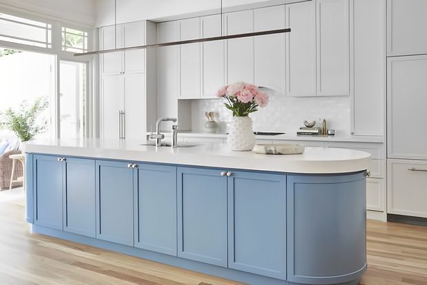 Bright, spacious kitchen featuring a large pale blue island with panelled cabinetry and rounded ends, white perimeter cabinetry, a white benchtop and splashback, integrated appliances, timber flooring, and a linear pendant light. Large windows and glass doors bring in abundant natural light.