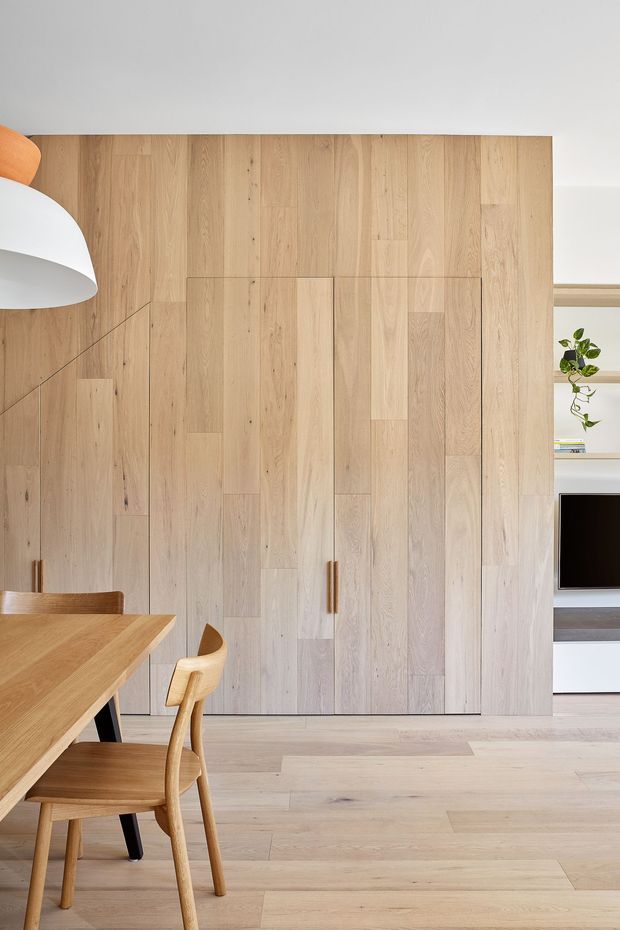 A bright, contemporary dining area featuring extensive light timber wall panelling with concealed storage doors and a slim vertical timber handle. Matching pale timber flooring and a simple timber dining table with curved wooden chairs create a cohesive warm-wood interior. A sculptural white pendant light hangs at left, while open shelving, a small plant, and a television are visible through the opening at right.