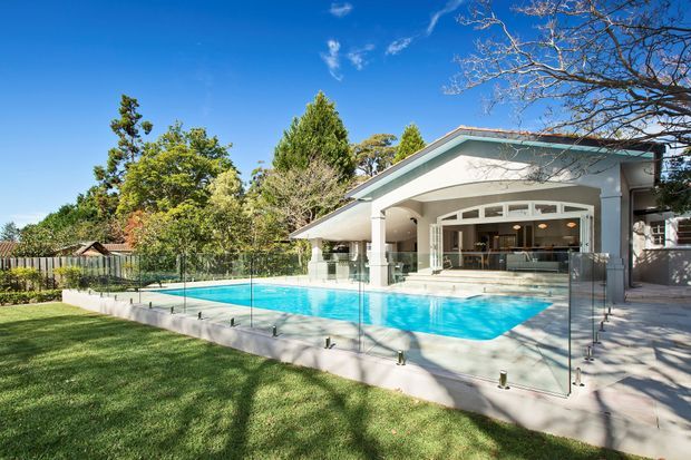 A bright exterior view of a modern single-storey home with a covered outdoor entertaining area opening onto a large swimming pool. The pool is enclosed by frameless glass balustrading, with a paved terrace, manicured lawn, mature trees and a clear blue sky. The house features a broad low-pitched roof, pale rendered walls, white columns and large glazed openings.