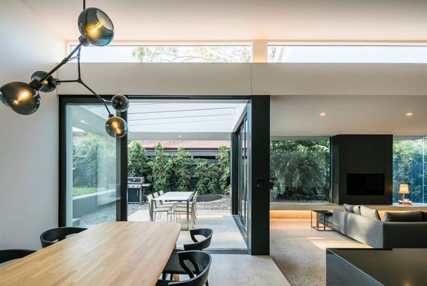 Open-plan contemporary dining and living area with a large light timber dining table, black dining chairs, sculptural multi-arm pendant lighting, dark-framed floor-to-ceiling glazing, and a living room with a built-in black fireplace feature. Glazing opens onto a landscaped courtyard, while high clerestory windows bring in additional daylight.