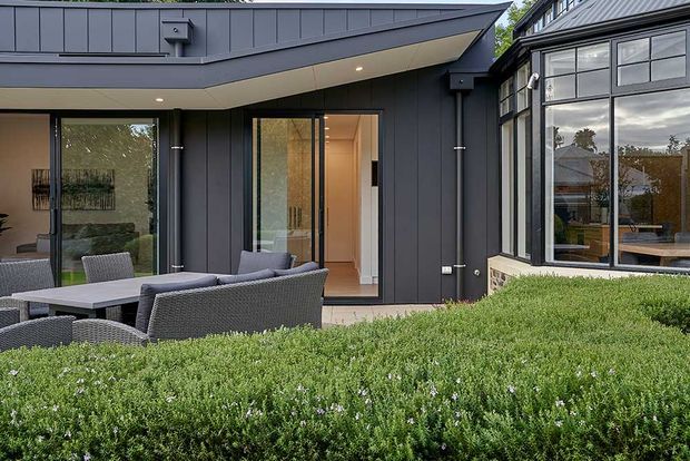 Contemporary residential extension viewed from a landscaped outdoor area, with dark vertical cladding, a low asymmetrical roof, large glazed openings, and an outdoor dining setting. A glazed, industrial-style conservatory or sunroom connects to the darker-clad portion of the home. Dense hedging occupies the foreground.