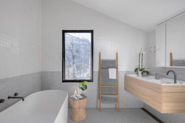 Bright contemporary bathroom with a freestanding white bathtub, wall-mounted timber vanity with integrated basin, black tapware, large mirrored cabinet, ladder-style timber towel rail, and a black-framed window. The walls and floor feature small-format light grey terrazzo-like tiles, with a darker grey horizontal wall band. Minimal styling and natural timber accents create a clean, calm atmosphere.