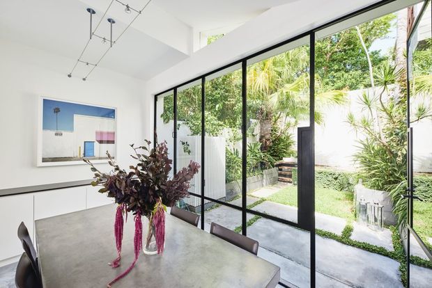 Bright contemporary dining area with a large concrete-finish dining table, sculptural pendant lighting, minimalist white built-in cabinetry, and full-height black-framed glazing overlooking a lush courtyard garden. A framed artwork and floral arrangement add colour to the otherwise restrained interior.