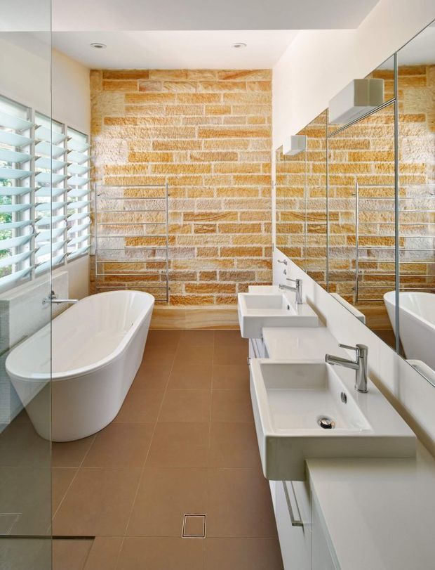 Bright contemporary bathroom featuring a freestanding white bathtub, double wall-mounted vanity basins, large mirrored surfaces, a frameless glass shower enclosure, and a distinctive exposed warm-toned brick feature wall. Timber-look louvred window shutters bring in natural light, while large matte brown floor tiles create a warm contrast.