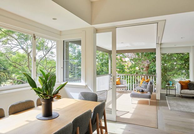 Bright open-plan dining and living area with large corner windows and folding glass doors opening onto a covered timber deck. The room has white walls and ceilings, light timber flooring, a light dining table with upholstered chairs, and a contemporary outdoor-facing lounge furnished in neutral tones. Lush greenery is visible through the glazing and around the deck.