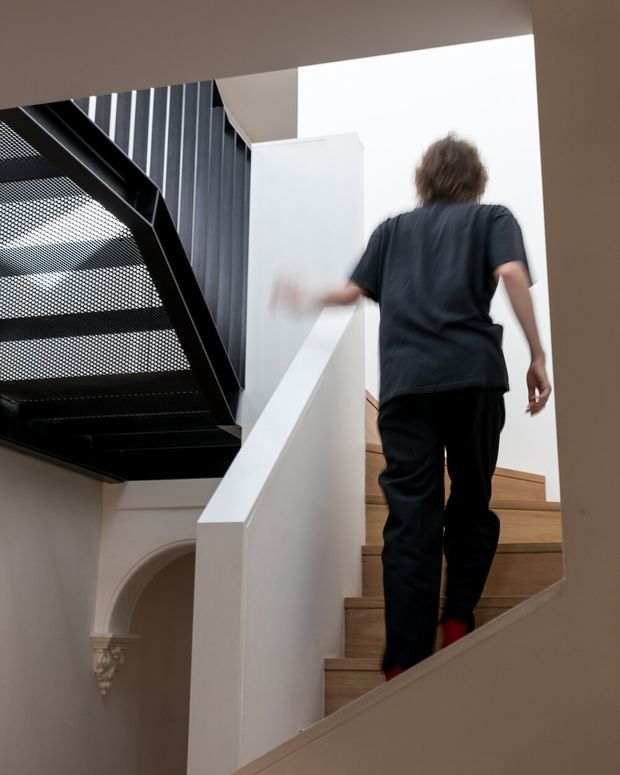 Interior architectural photograph looking up a staircase, with a person ascending light timber steps. The stair features a crisp white balustrade and wall surfaces, alongside a contrasting black perforated metal stair or landing structure. An arched architectural opening and decorative moulding are visible below, creating a contemporary intervention within a more traditional shell.