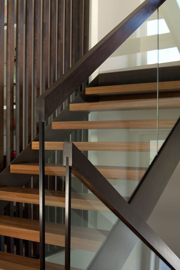 Close-up interior view of a contemporary open-riser staircase. Warm timber treads are supported by dark metal framing, with dark timber handrails and clear glass balustrades. Vertical dark timber slats form a feature wall alongside the stair, while bright glazing and a high stairwell create an airy, refined composition.