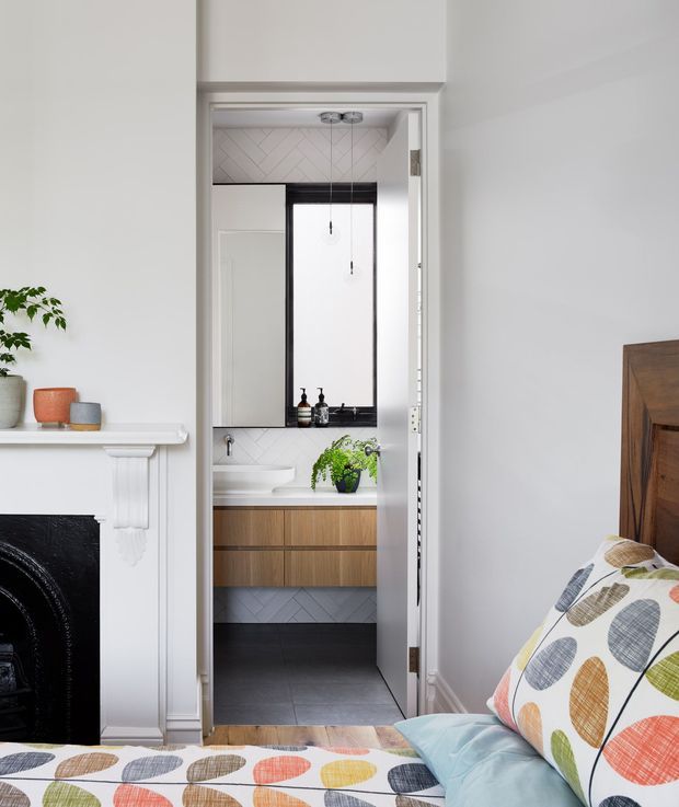 A bright bedroom viewed toward an open bathroom doorway. The bedroom has white walls, a decorative traditional fireplace surround with black firebox, a wooden bed frame, and colourful patterned bedding. Through the doorway is a compact contemporary bathroom with a floating timber vanity, white basin, large mirror, dark-framed window, pale herringbone wall tiles, and dark floor tiles. A small potted plant adds greenery.