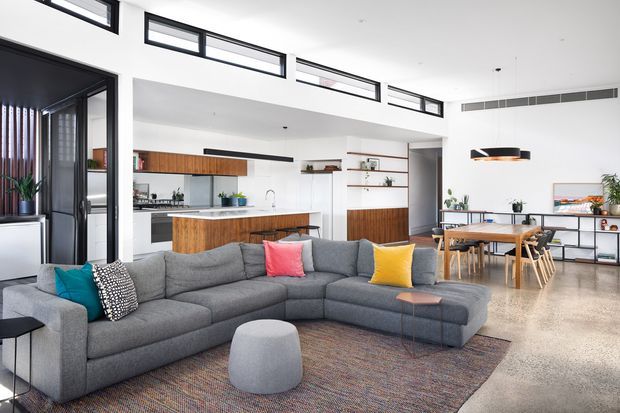 Open-plan contemporary living, dining and kitchen area with a large grey sectional sofa in the foreground, timber kitchen island and cabinetry, dining table, built-in shelving, polished concrete flooring, and abundant high-level glazing. The bright interior combines white surfaces, warm timber, black-framed glazing, and colourful cushions.