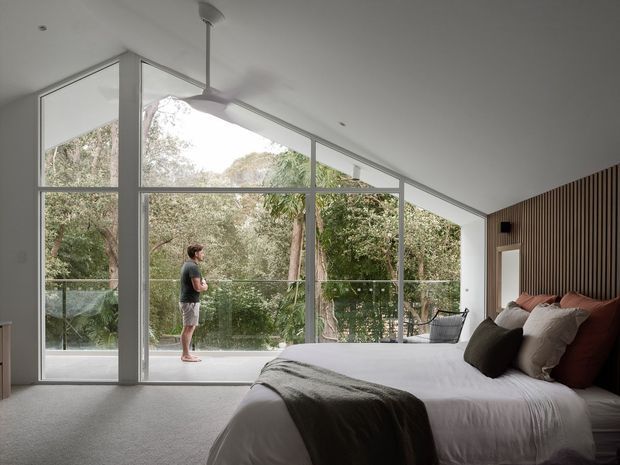 A contemporary bedroom with a large bed in the foreground, a dark vertical timber-slat feature wall behind the bed, and expansive floor-to-ceiling glazing opening onto a balcony or terrace surrounded by trees. The room has a sloping ceiling, soft neutral furnishings, carpeted flooring, and a ceiling fan. A person stands outside on the glazed terrace.