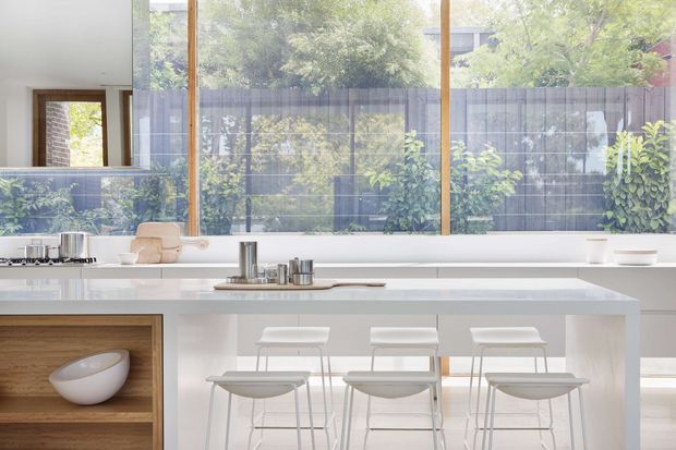 Bright, airy contemporary kitchen with a long white island and integrated seating for four. The space features white cabinetry and benchtops, a timber-framed full-height glazed wall overlooking a planted courtyard, timber open shelving, and minimal white stools. Natural light and greenery create a clean indoor-outdoor connection.