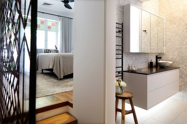 A bright, contemporary bathroom dominates the foreground, featuring a floating white vanity with a dark benchtop, vessel basin, mirrored cabinet, black fixtures, and a wall clad in small hexagonal tiles. A black metal-framed shower screen and ladder-style towel rail are visible. Through the opening beside the bathroom, a bedroom with a bed, rug, curtains, and daylight is visible, separated by a short timber step-up and decorative dark lattice screen.