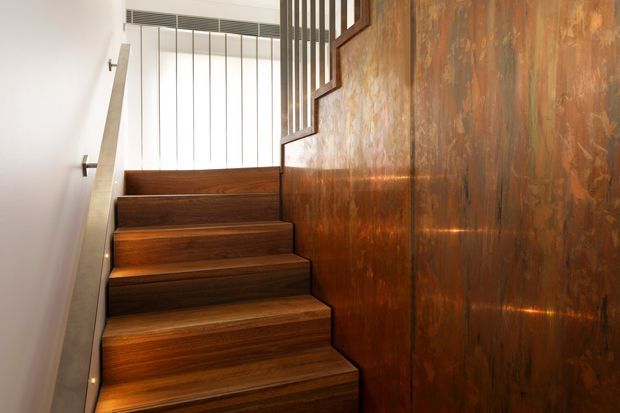 Interior view of a modern stairway with broad dark timber treads and risers, a warm patinated copper or bronze-toned wall along the right side, and slim vertical metal balusters forming a guardrail at the upper landing. A simple pale handrail and white wall contrast with the rich, reflective finishes. Integrated step lighting adds a warm glow.