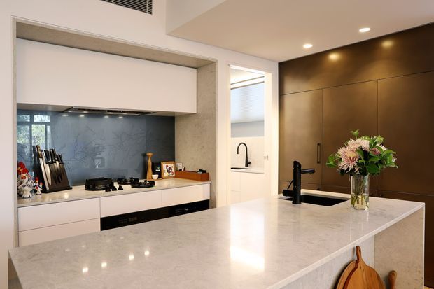 A modern contemporary kitchen featuring a large pale stone island with an integrated sink and matte black tap, white handleless cabinetry, a dark stone or glass splashback, and a full-height dark timber-toned cabinetry wall. Recessed ceiling lights, concealed rangehood cabinetry, and a partially visible adjacent utility or pantry area are also shown.