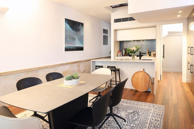 Bright contemporary open-plan dining and kitchen interior. A large rectangular dining table with black and white chairs occupies the foreground, while a compact kitchen with a pale stone island, integrated cabinetry, tiled splashback and timber flooring sits beyond. White walls, recessed lighting, artwork, a patterned rug and warm wood tones create a clean, inviting space.